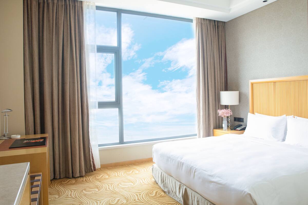 Habitación de hotel con un ventanal grande, una cama, una mesita de noche con lámpara y un escritorio con silla.