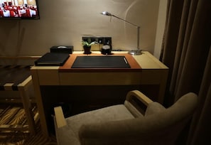 Quarto de hotel com escrivaninha, cadeira, televisão, abajur e planta em vaso.
