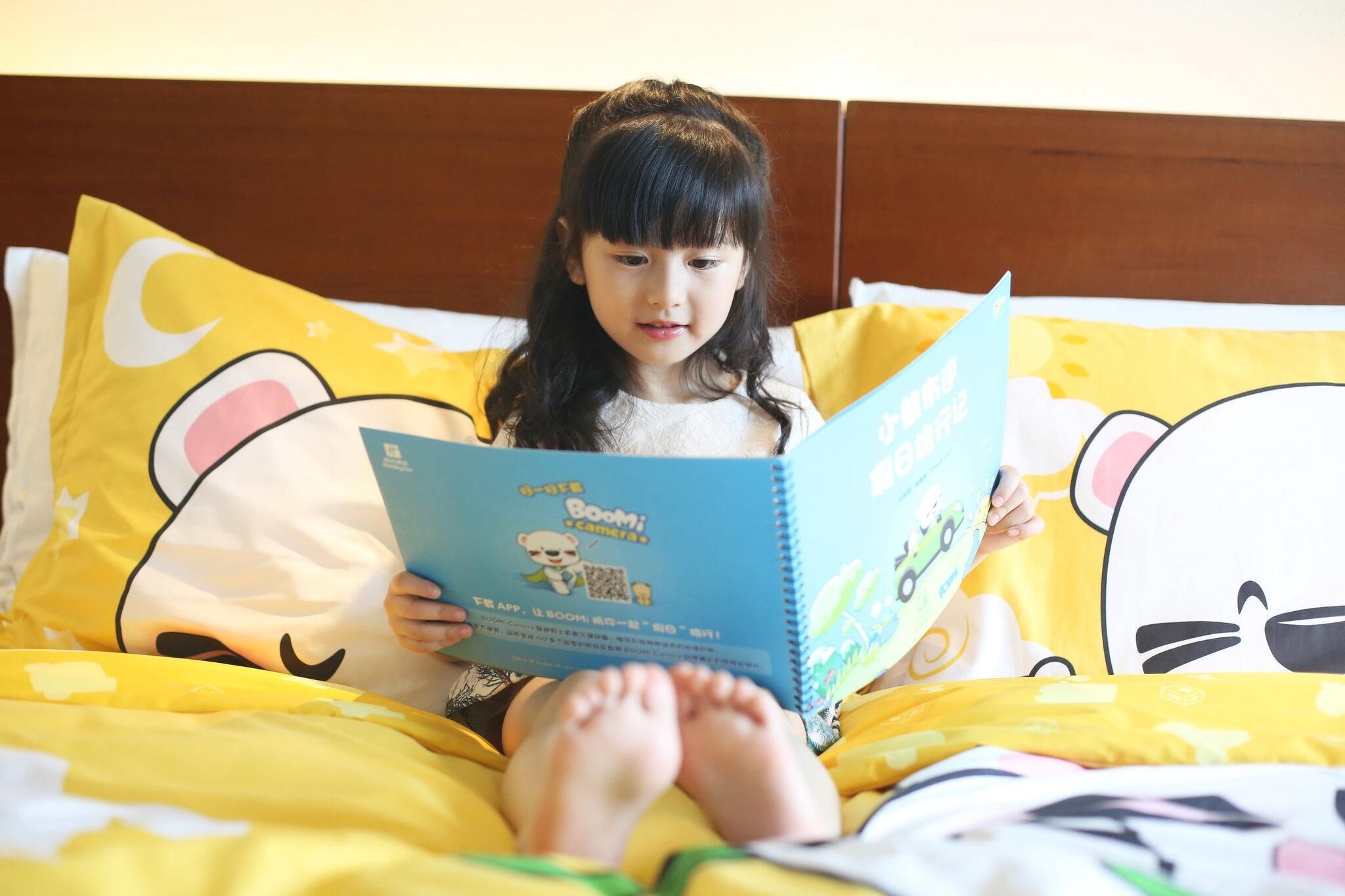 A child sitting on a bed with yellow bedding and cartoon-themed pillows, reading a book.
