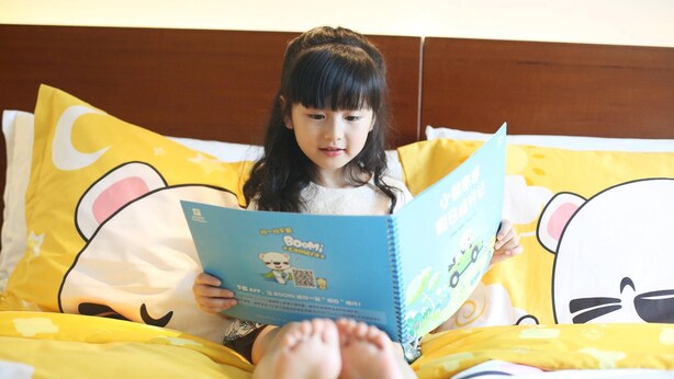 Un enfant est assis sur un lit dont la literie est jaune et les oreillers sont décorés de motifs de dessins animés, et il est en train de lire un livre.