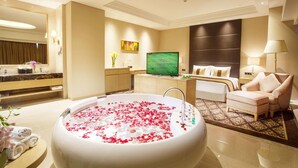 Une chambre d’hôtel dotée d’une grande baignoire remplie d’eau et de pétales de rose, d’une télévision, d’un lit, d’un canapé et d’une salle de bain avec un miroir et un meuble-lavabo.