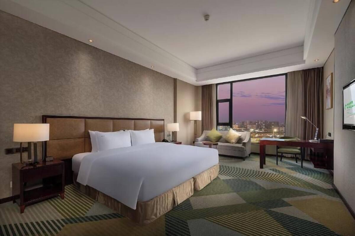 Habitación de hotel con una cama grande, un escritorio, una silla y vistas de la ciudad al anochecer.