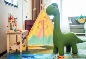 Uma área de recreação infantil com um brinquedo de dinossauro verde, uma mesa de brincar de madeira e uma tenda colorida.