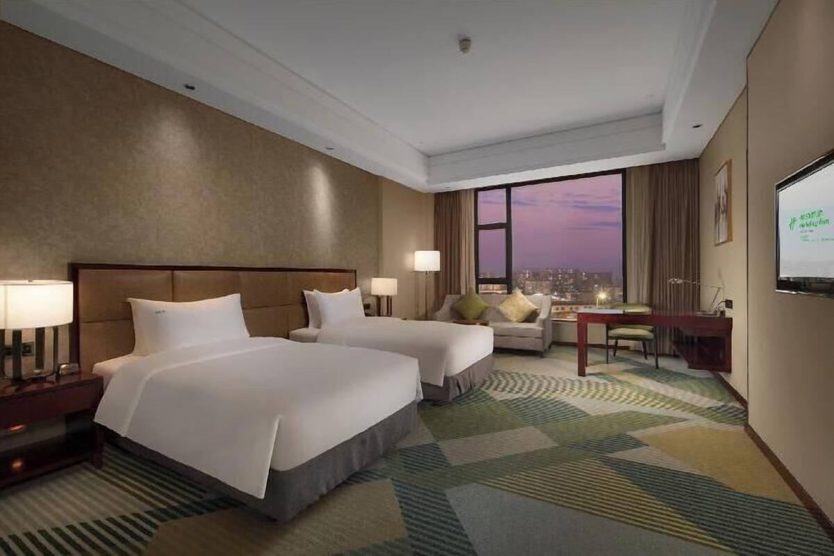 Habitación de hotel con dos camas, un escritorio, una silla, un televisor y un ventanal con vistas a la ciudad.