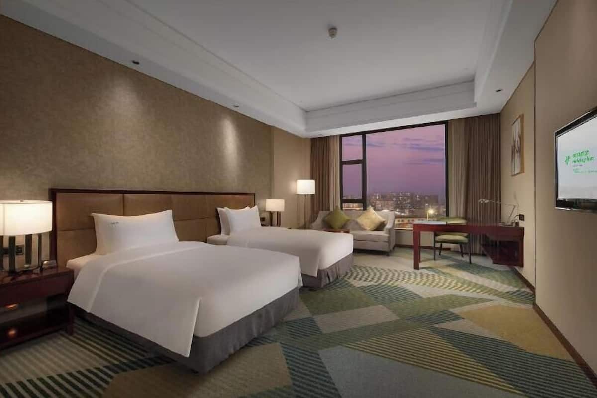 Quarto de hotel com duas camas, uma escrivaninha, uma cadeira, uma televisão e uma janela grande com vista para a cidade.