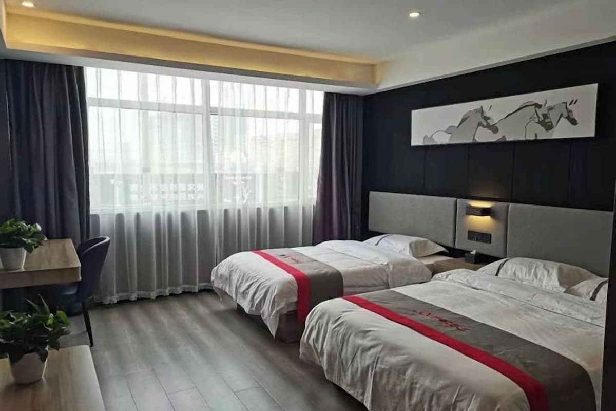 Habitación de hotel con dos camas, un escritorio, una silla, una planta en maceta y un amplio ventanal con cortinas.