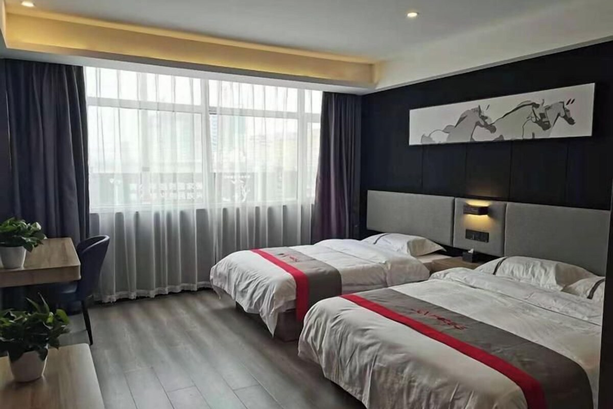 A hotel room with two beds, a desk, a chair, a potted plant, and a large window with curtains.