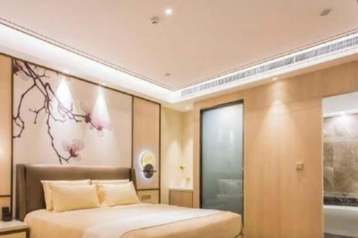 A modern hotel room with a large bed, a bedside table, a wall-mounted lamp, and a decorative wall piece.