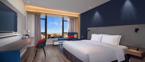 A modern hotel room with a large bed, a desk, a chair, and a view of the city.