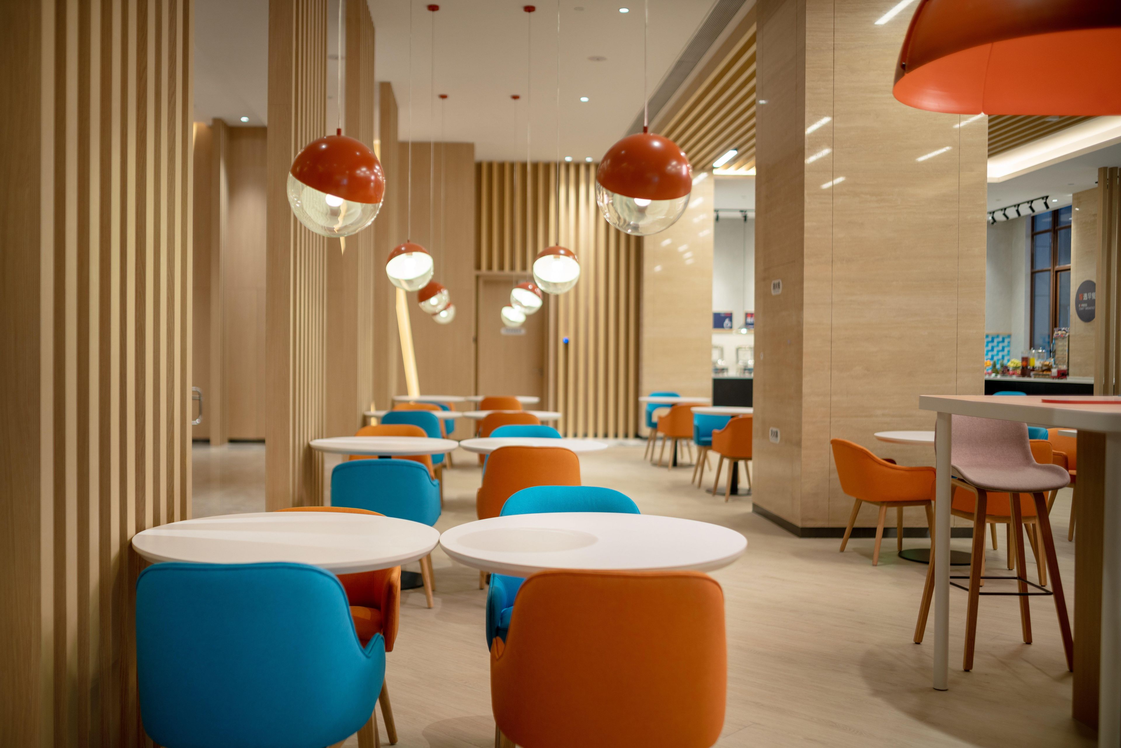 A modern dining area with round tables and colorful chairs, featuring a wooden panel wall and hanging pendant lights.