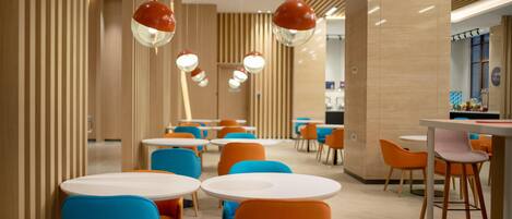 A modern dining area with round tables and colorful chairs, featuring a wooden panel wall and hanging pendant lights.