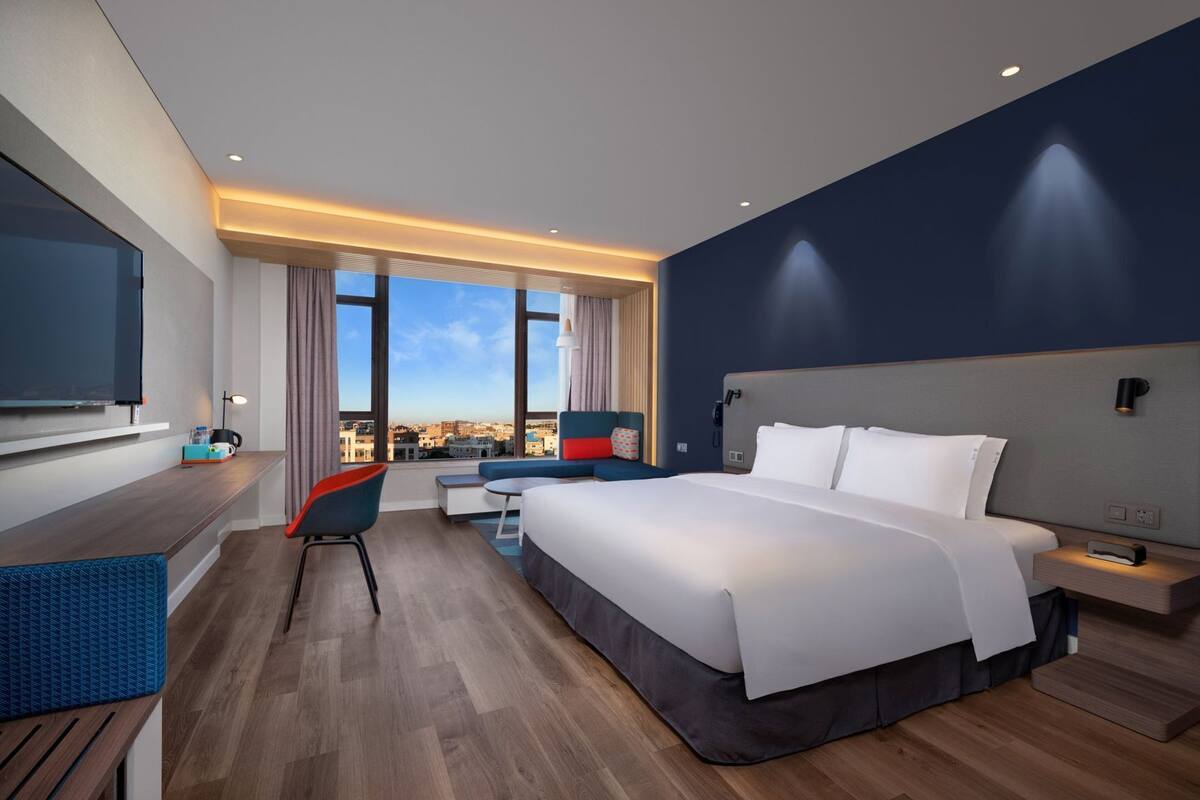 Habitación de hotel moderna con una cama grande, un escritorio, una silla, un televisor y vistas a la ciudad.