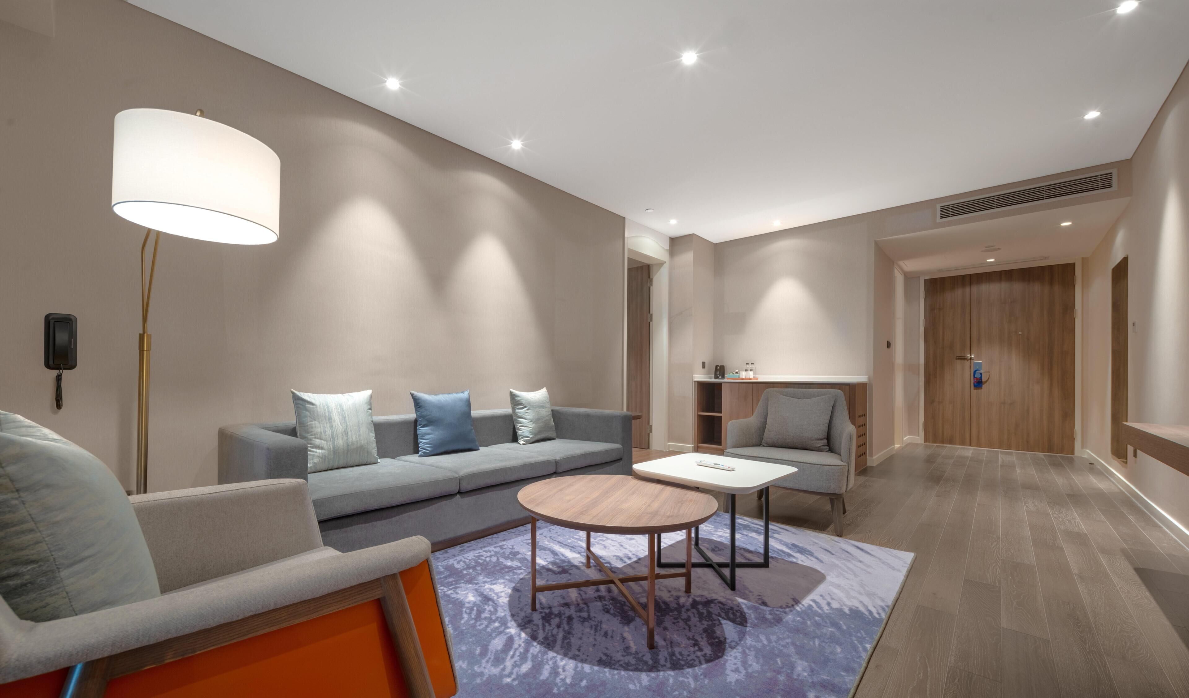 A modern hotel room with a sofa, armchair, round coffee table, and a lamp.