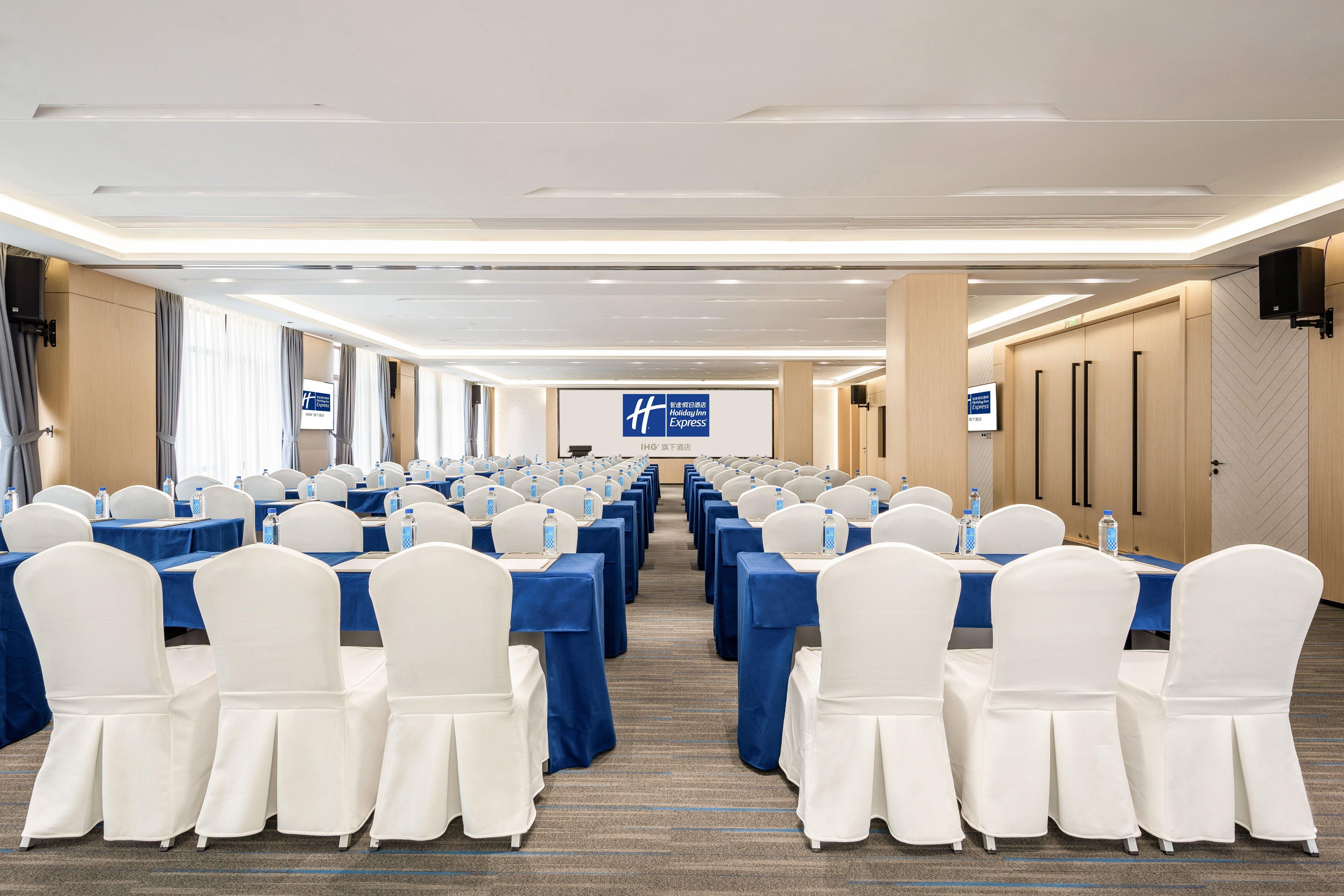 Un salón de conferencias con filas de mesas y sillas, una pantalla grande al frente y un letrero que indica que forma parte de la cadena Holiday Inn Express.