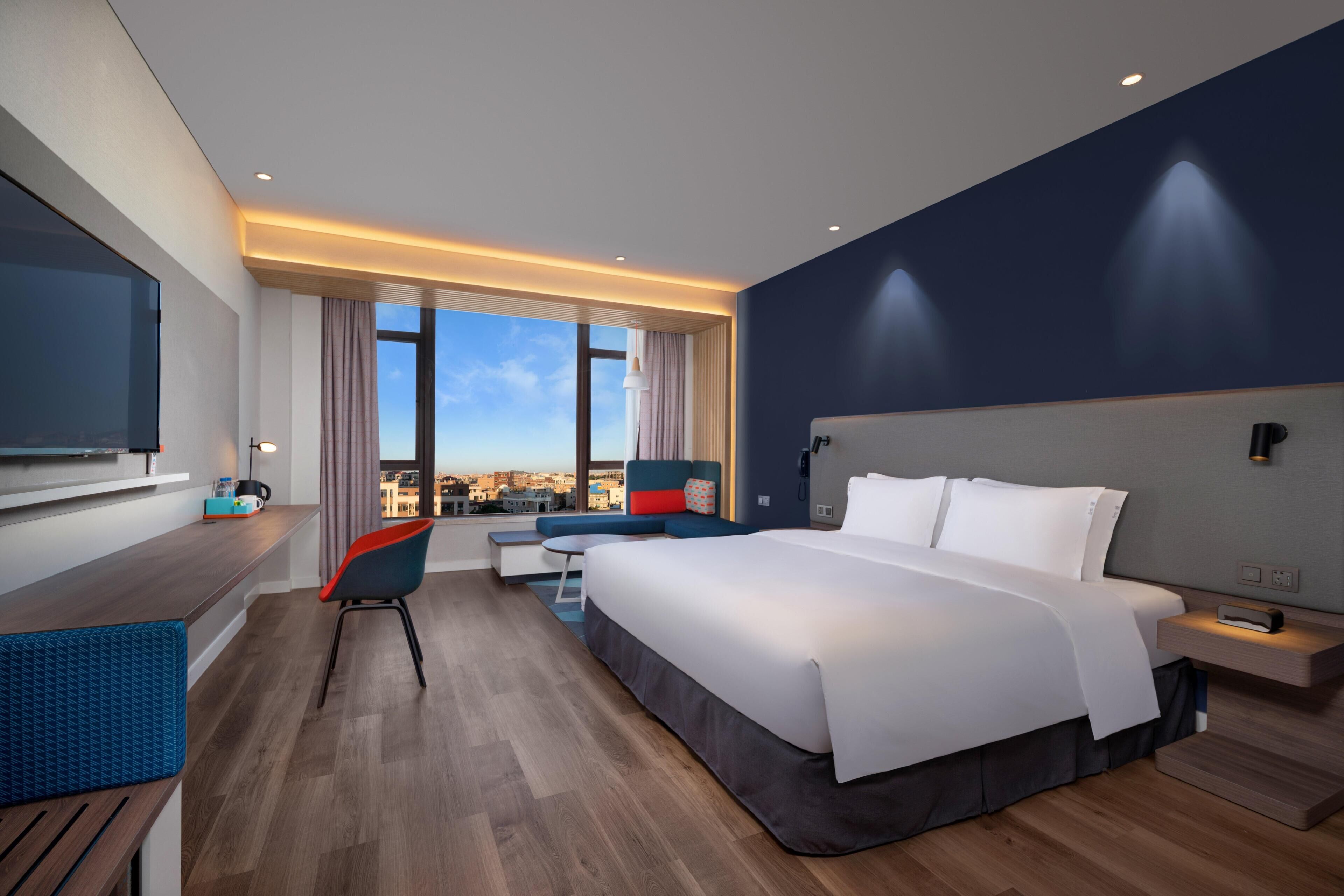 A modern hotel room with a large bed, a desk, a chair, and a view of the city.