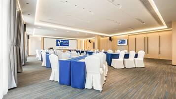 Una sala de conferencias con filas de mesas y sillas, una pantalla de proyector y un cartel con el nombre del hotel.