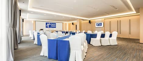 Una sala de conferencias con filas de mesas y sillas, una pantalla de proyector y un cartel con el nombre del hotel.