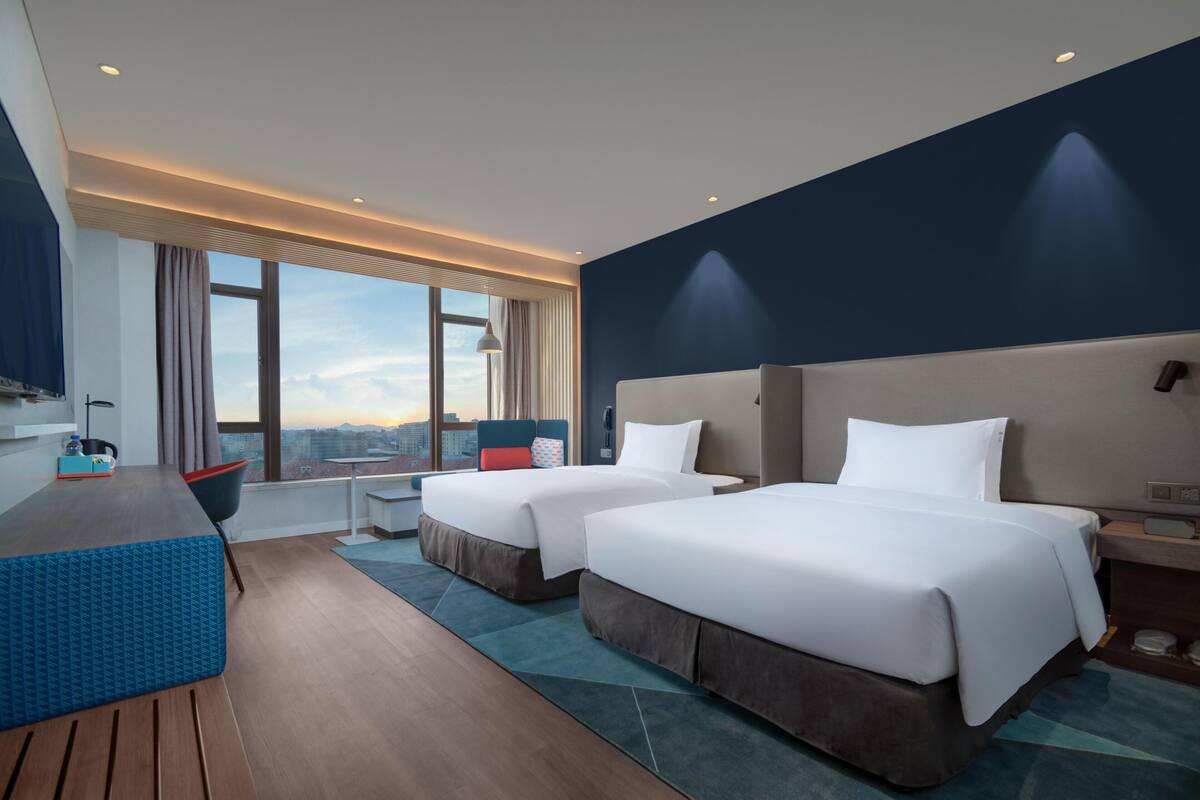 Habitación de hotel con dos camas, un escritorio, una silla y un ventanal con vistas a la ciudad.
