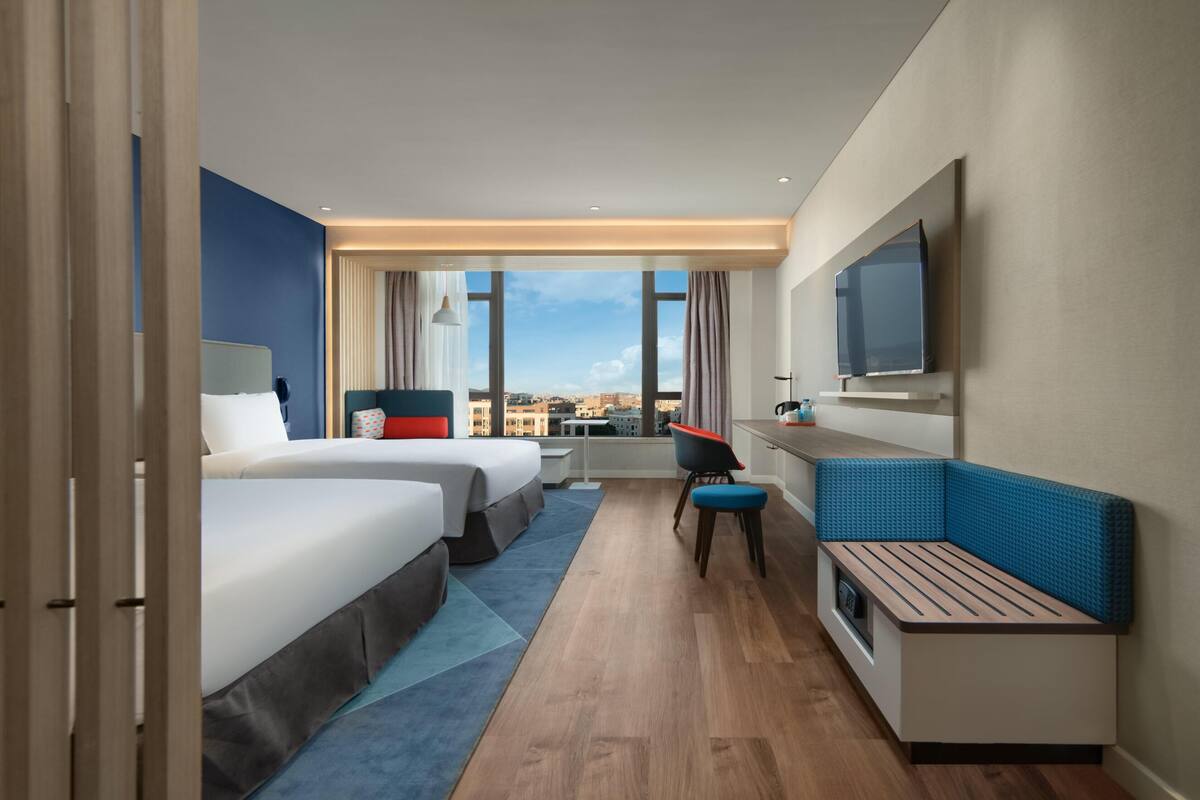 Habitación de hotel con dos camas, un escritorio, un televisor y vistas a la ciudad.