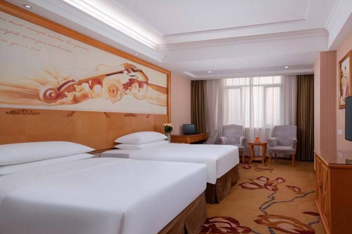 A hotel room with two beds, a desk, a chair, a television, and a large floral wall decoration.