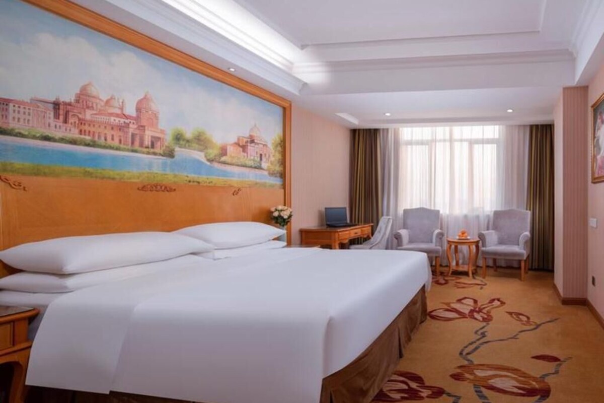 A hotel room with a large bed, a desk, two chairs, a laptop, and a painting of a building on the wall.