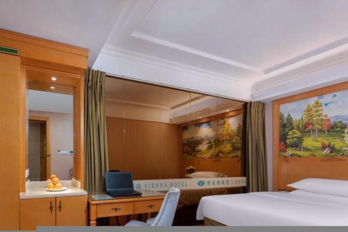 A hotel room with a bed, a desk with a laptop, a mirror, and a painting on the wall.