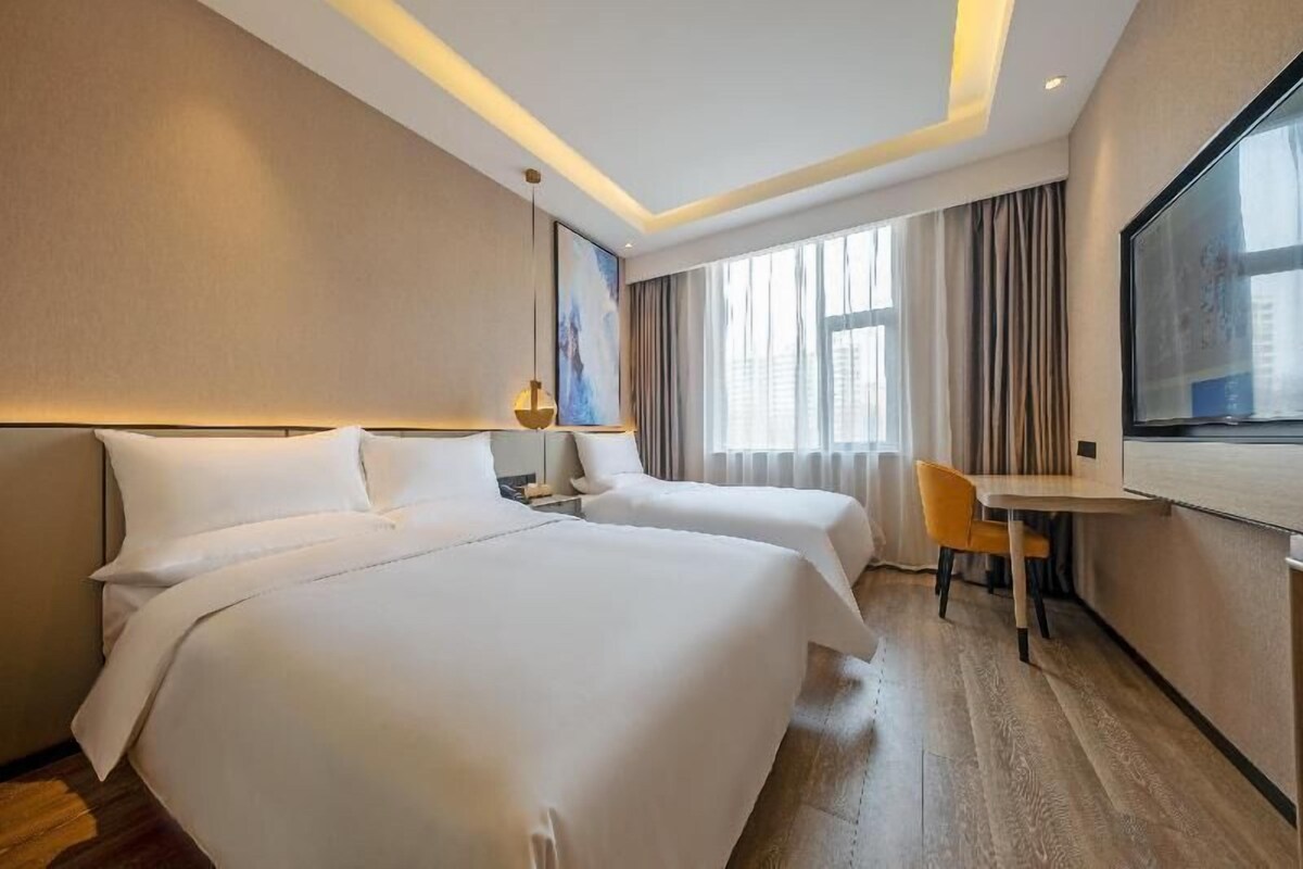 A hotel room with two beds, a desk, a chair, a large window, and a painting on the wall.