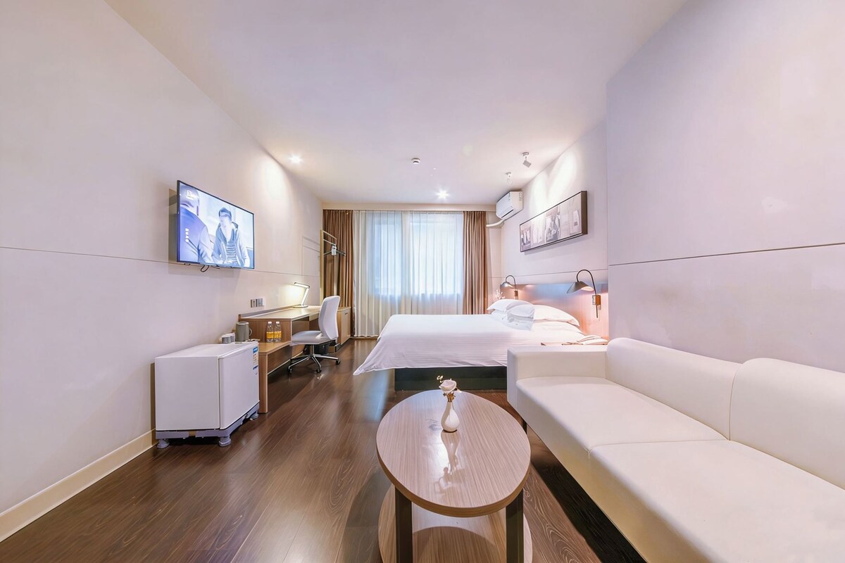 A hotel room with a bed, a sofa, a desk, a chair, a TV, and a small table with a vase.