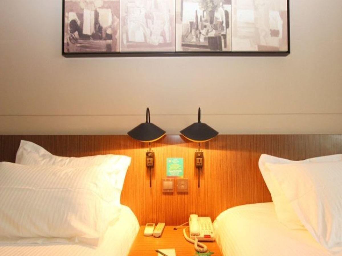 Quarto de hotel com duas camas, cabeceira de madeira, duas luminárias fixadas na parede, um telefone e um quadro emoldurado sobre as camas.