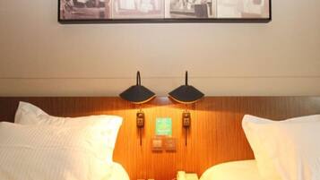 Quarto de hotel com duas camas, cabeceira de madeira, duas luminárias fixadas na parede, um telefone e um quadro emoldurado sobre as camas.