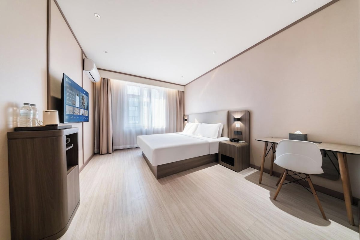 A hotel room with a bed, bedside table, desk, chair, and a TV on the wall.
