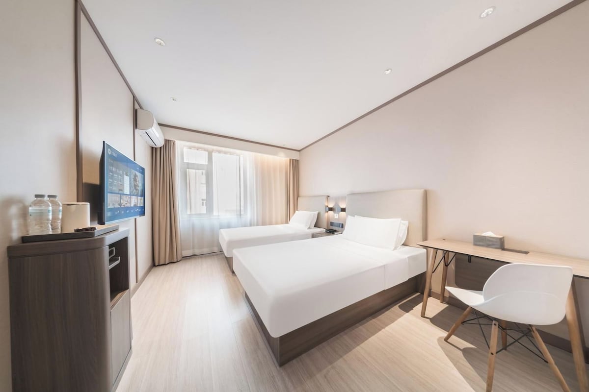 A modern hotel room with a large bed, a desk, a chair, a TV, and a minibar.