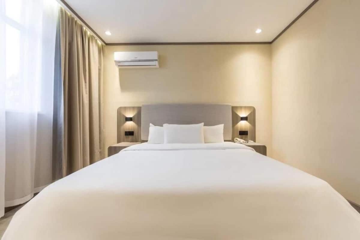 A hotel room with a large bed, two bedside tables with lamps, an air conditioning unit, and a window with curtains.
