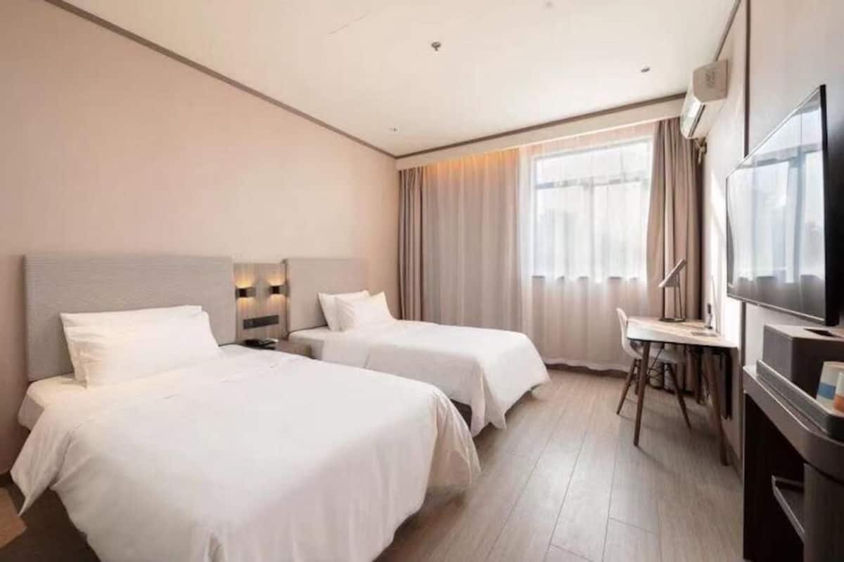 A hotel room with two beds, a desk, a TV, and a large window with curtains.