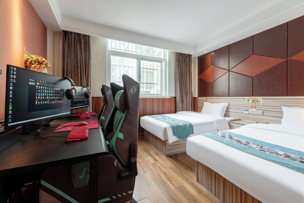 A hotel room with a desk, computer, gaming chair, two beds, and a window.