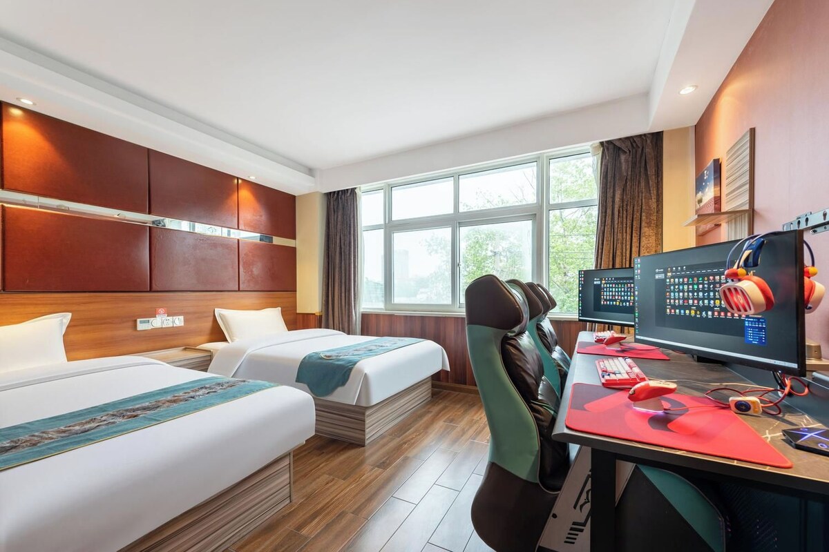 A hotel room with two beds, a desk with a computer and headphones, and a large window.