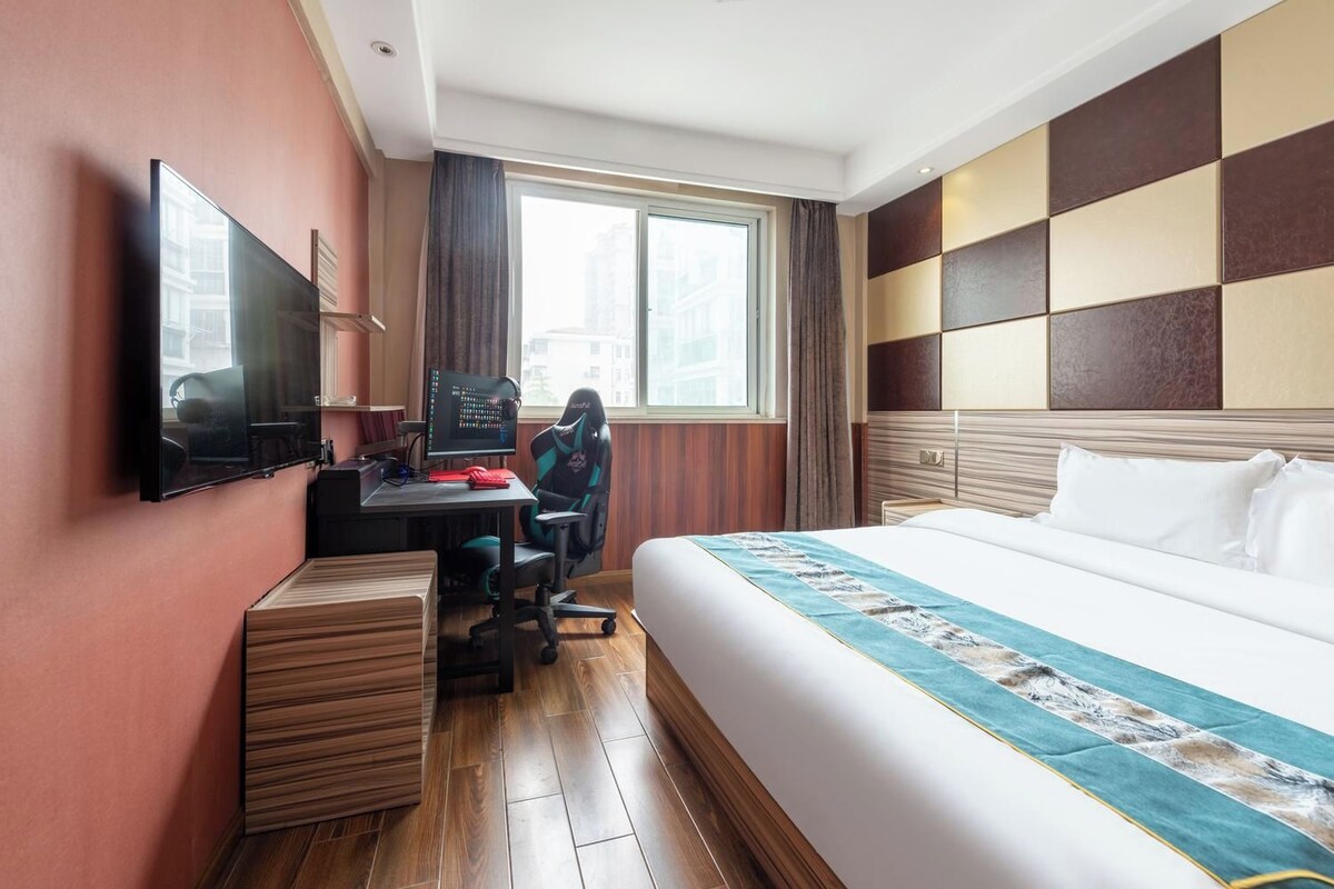 A hotel room with a bed, a desk with a computer, a chair, and a TV mounted on the wall.