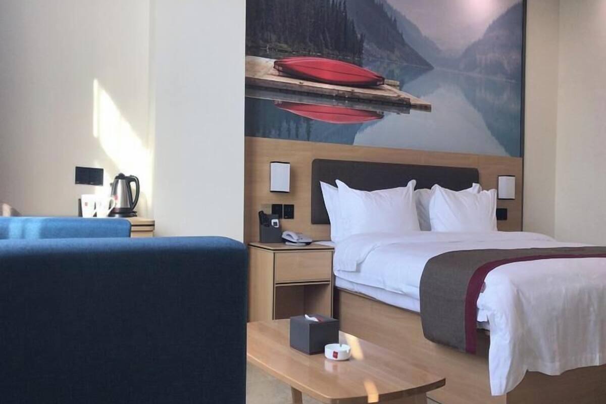 Et hotelværelse med seng, skrivebord, stol, natbord, kaffemaskine og et maleri af et bjerglandskab.