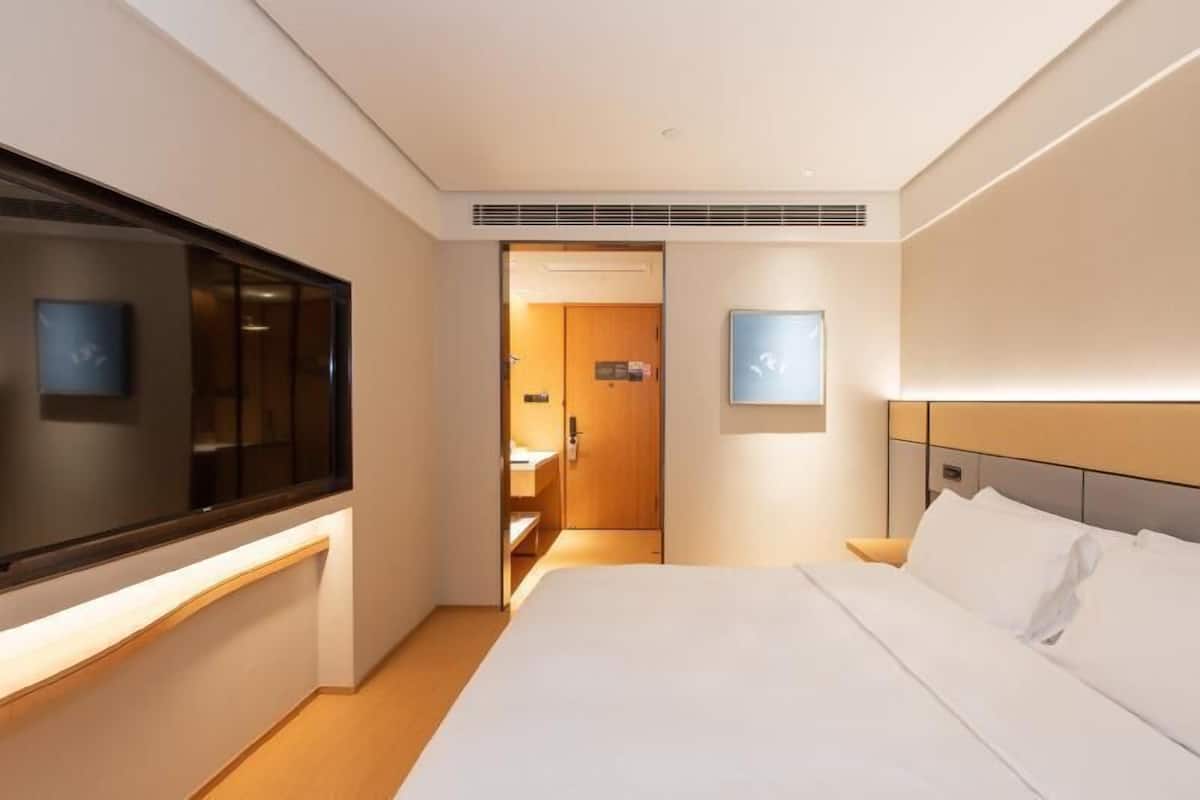 A hotel room with a bed, a television, a door leading to a bathroom, and a wall-mounted light fixture.