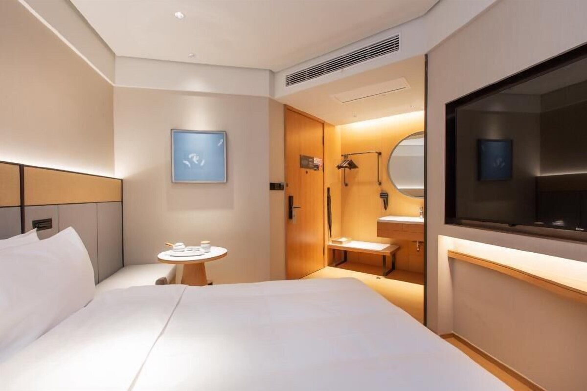 Una habitación de hotel moderna con cama, mesita de noche, una pequeña mesa redonda, un espejo y televisión.