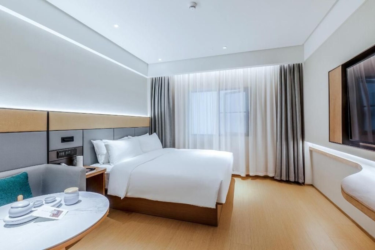 Habitació d'hotel moderna amb un llit gran, una tauleta de nit, una taula rodona amb una cadira i una finestra ampla amb cortines.
