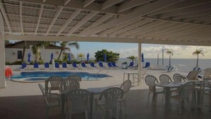 Zona de piscina exterior con sillones y mesas, un área de estar cubierta y vista al océano.