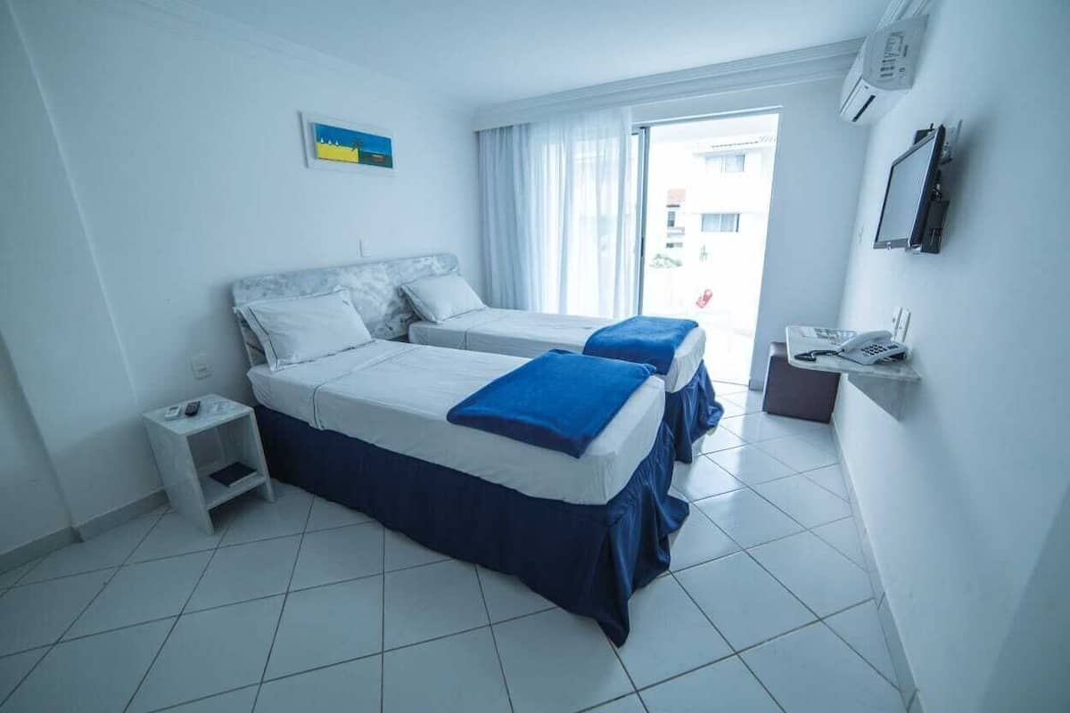 Ein Hotelzimmer mit zwei Betten, einem kleinen Nachttisch, einem Schreibtisch mit Telefon und einem Fenster mit Vorhängen.