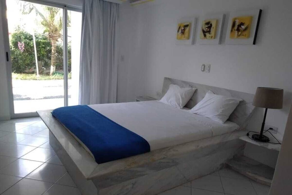 Ein Schlafzimmer mit einem Bett, einem Nachttisch, einer Lampe und Blick ins Freie.
