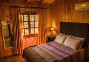 Quarto de madeira com cama, janela com cortinas, mesa de cabeceira e ar-condicionado fixado na parede.