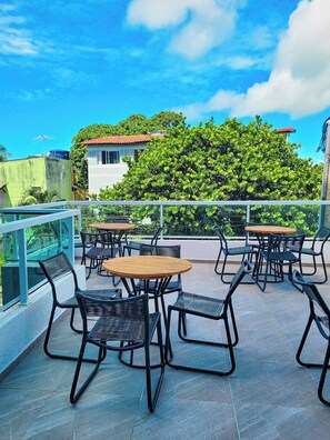 A rooftop patio with outdoor seating, a view of a building with a red roof, and a large green bush. - Porto Sol Vila By Aft (Ipojuca)