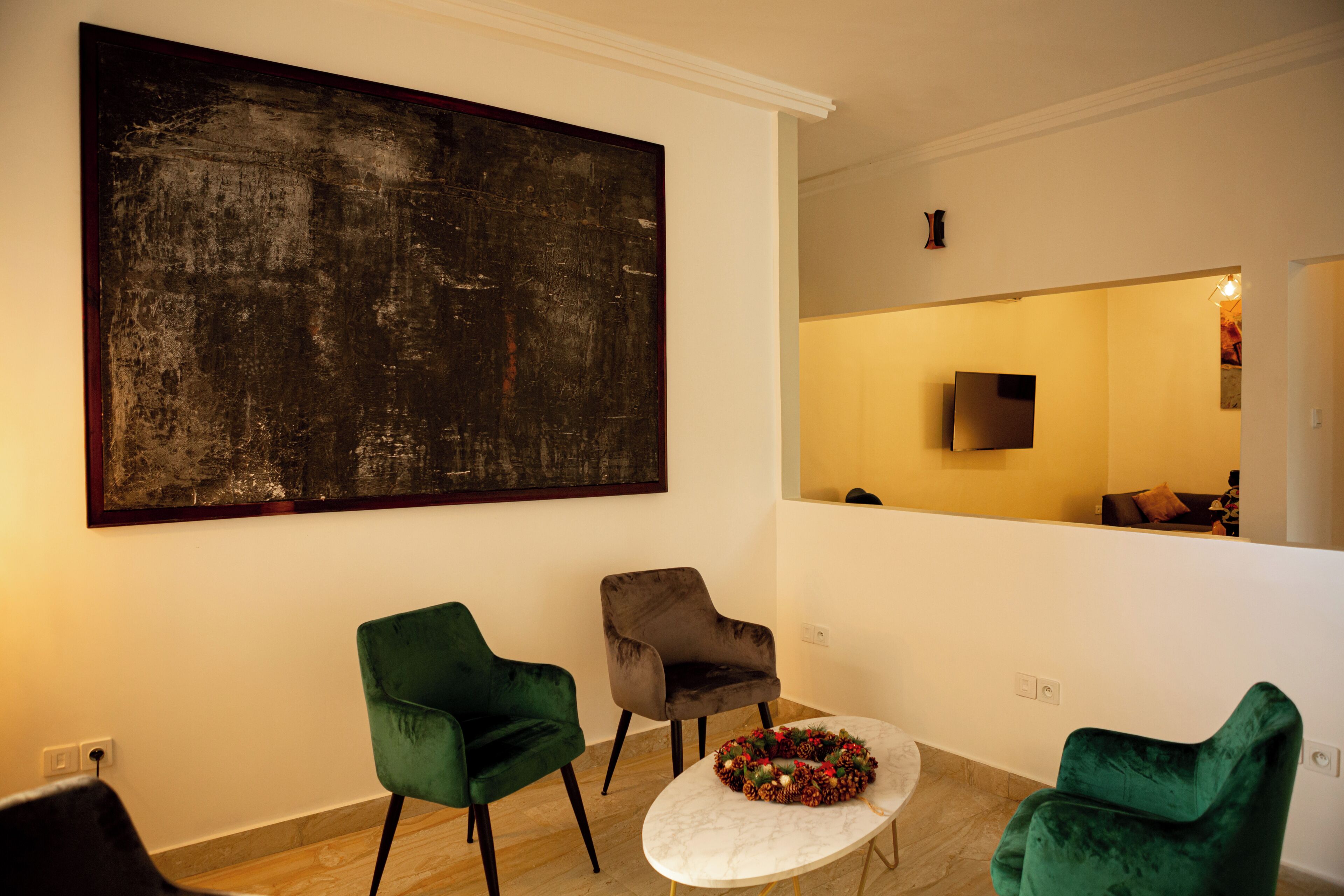 Una habitación con un cuadro abstracto grande, dos sillas verdes, una mesa redonda pequeña con un arreglo floral, y un televisor al fondo.