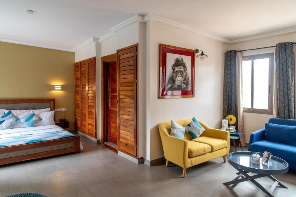 Habitación de hotel con cama, sofá amarillo, sofá azul, mesa redonda y un cuadro en la pared.