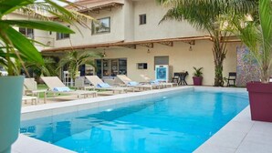 A swimming pool with lounge chairs, palm trees, and a building with a tiled roof. - LE KARE EBENE (Cotonou)