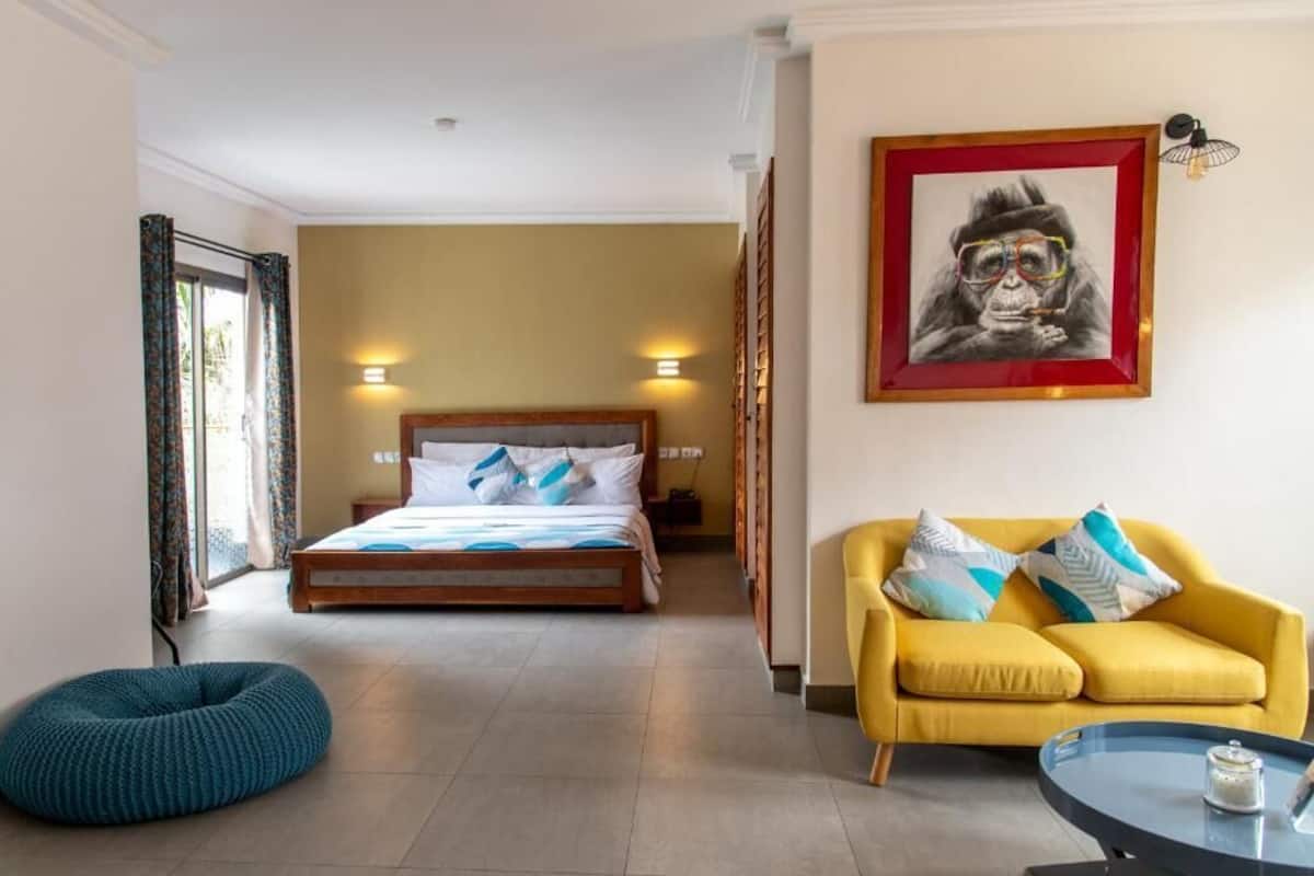 Habitación de hotel con cama, sofá amarillo, sillón de peluche azul y un cuadro en la pared.